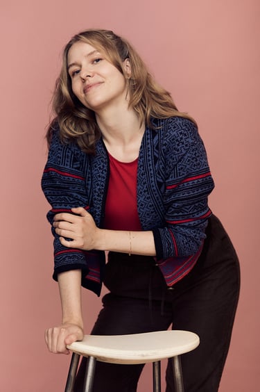 A young woman wearing a blue sweater and red t-shirt leaning on a stool.
