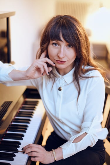A woman sitting by a piano in a white blouse.