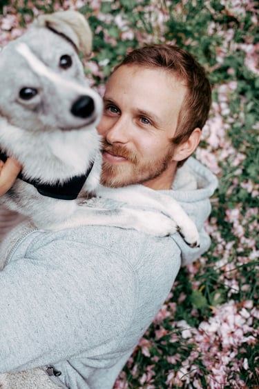A portrait of a man lying on grass with pink petals looking at his dog.