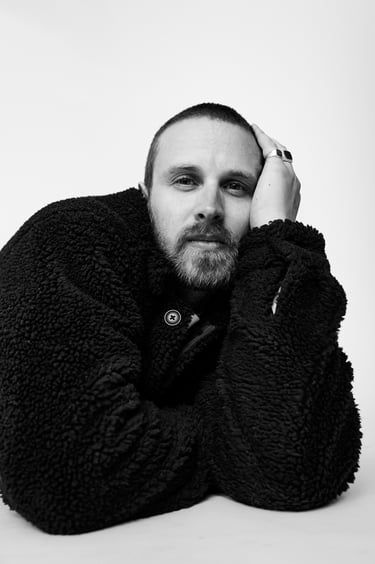 A black and white portrait of a man lying on a floor in a black woollen jacket.