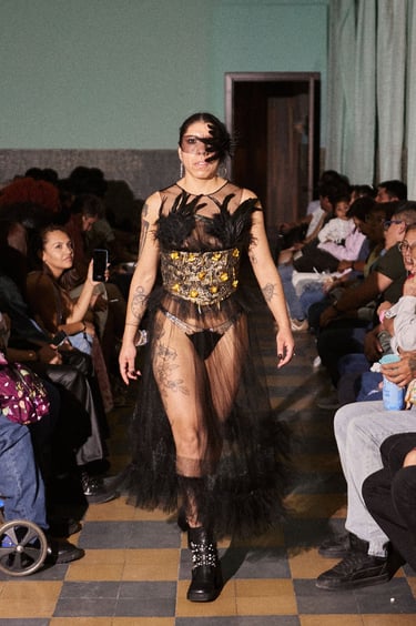 A model walks a fashion runway wearing a bold red structural top and a sheer tulle skirt.