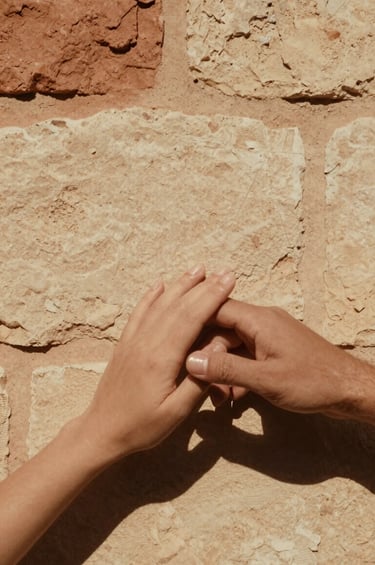 A cinematic, vertical frame capturing a couple's hands gently touching against a background of a sun-bleached stone wall. The style is minimalist and emotive, focusing on textures. The warm sandy tones of #FDF8F0 dominate the scene, with deep terracotta #AD7B5B shadows providing depth and an authentic, timeless feel.
