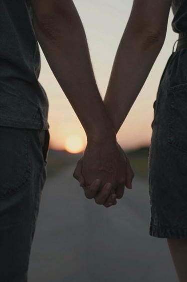 A close-up, cinematic detail shot of two people holding hands during a warm sunset stroll. Focus is on the authentic interaction and natural beauty. The color palette emphasizes rich Charcoal #2C2C2C textures in the background and warm sun-drenched skin tones.