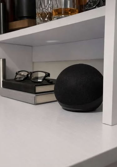  Compact smart speaker resting on a white shelf beside stacked books, glasses, and home decor