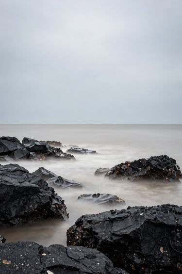 A fine art photograph of the Brest coastline, captured with a long exposure to make the water appear like a soft off-white mist. The rocks are a deep dark charcoal, and the sky is a pale, calm grey. The composition is clean, centered, and minimalist.