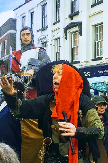 Medieval reenactors in costume including a knight and jester at a crowded outdoor heritage street market.