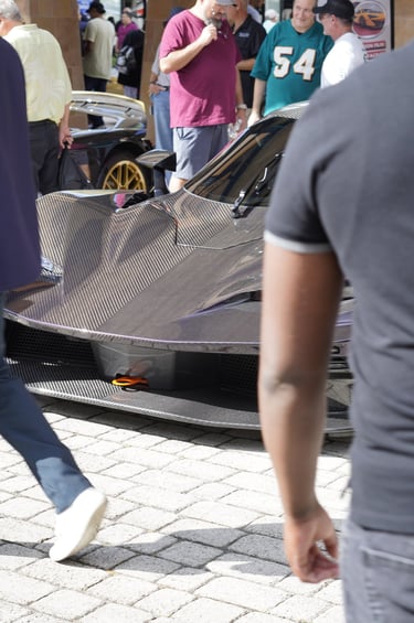 A purple exposed carbon fiber hypercar on display at an outdoor luxury auto show with spectators.