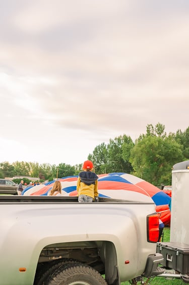 A child in a pickup truck watching a colorful hot air balloon inflate at a festival.