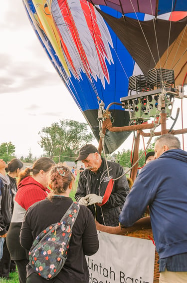 A hot air balloon pilot demonstrates burner equipment to a group of people at a balloon festival.