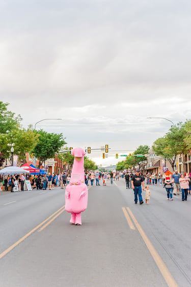 A large pink dinosaur mascot walks down a crowded city street during a summer community festival.