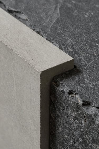 A detailed close-up photograph of architectural materials. A slab of light neutral grey concrete meets a textured dark slate grey stone. The lighting is soft and directional, focusing on the tectonic precision and the low-impact materials used by Estúdio Roda.