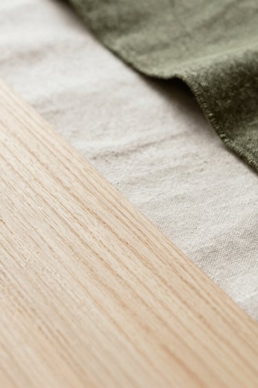 A macro photograph of natural textures used in sustainable design, such as light-toned wood grain and organic linen fabric. The composition is artistic and minimalist, shot in a bright, airy environment in South America. The palette focuses on off-white and muted olive green.