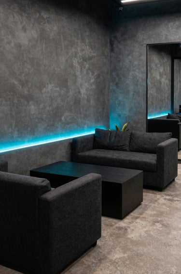 A stylish and modern lounge area in a South American / Colombian creative studio, deep charcoal black furniture with electric neon cyan lighting strips, gunmetal grey walls, professional and cinematic urban vibe.