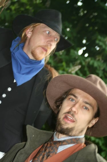 Two men in vintage Western outlaw costumes wearing cowboy hats and neckerchiefs outdoors.