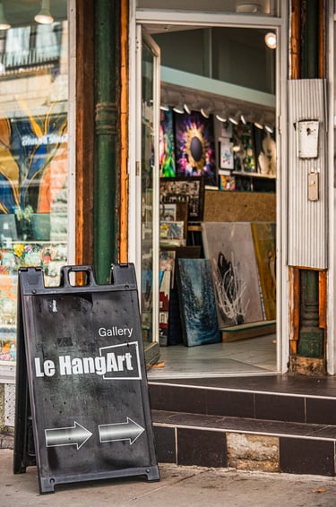 A sandwich board on the sidewalk, pointing to the entry of an art gallery with paintings inside