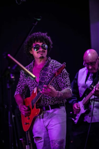A guitarist with curly hair and sunglasses performs live music on a red electric guitar at a concert venue.