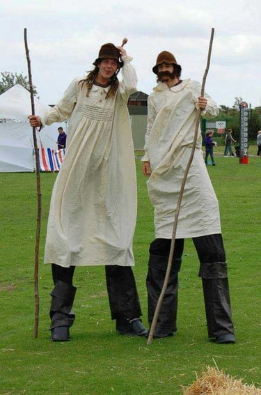 Two street performers on stilts wearing traditional smocks and hats at an outdoor festival.