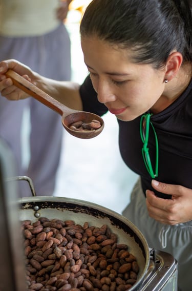 A woman smells roasted cacao beans in a wooden spoon over a large pan during traditional chocolate production.