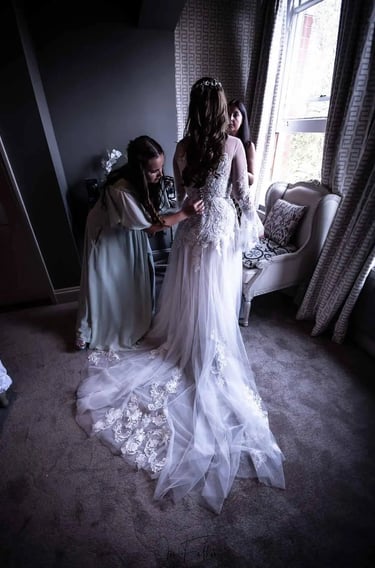 Bridesmaid helping a bride into her lace wedding dress with a long train by a window.