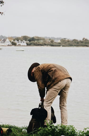 A man in a brown waxed jacket and hat packing a backpack by a scenic lake or river.