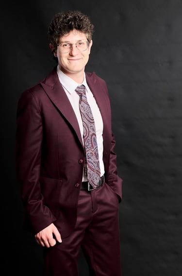 Professional portrait of a man in a maroon suit and paisley tie against a black background.