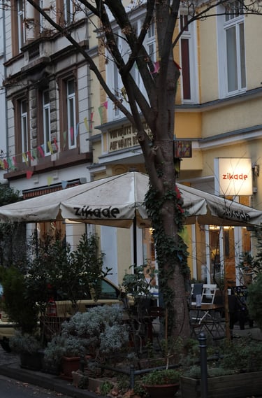Charming Zikade cafe storefront in Colognewith outdoor seating, patio umbrellas, and colorful flags.