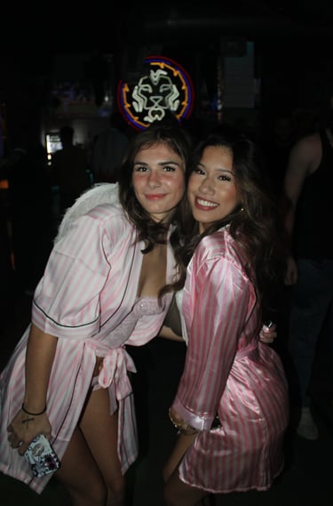 Two smiling women wearing pink striped satin robes and angel wings at a nightclub party.