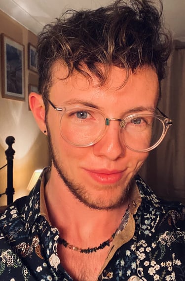 White trans masculine person with blue eyes, glasses, light brown curls and facial hair, smiling