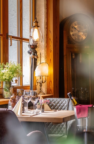 Eleganter Restauranttisch mit Weingläsern am Fenster, antiker Standuhr und warmer Lichtstimmung.
