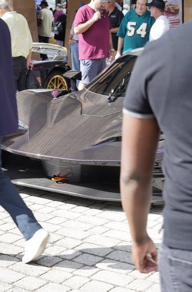 A purple exposed carbon fiber hypercar on display at an outdoor luxury auto show with spectators.