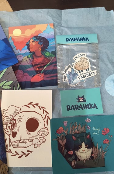 A collection of Rarainka art prints and stickers featuring a black cat, floral skull, and diverse character illustrations.