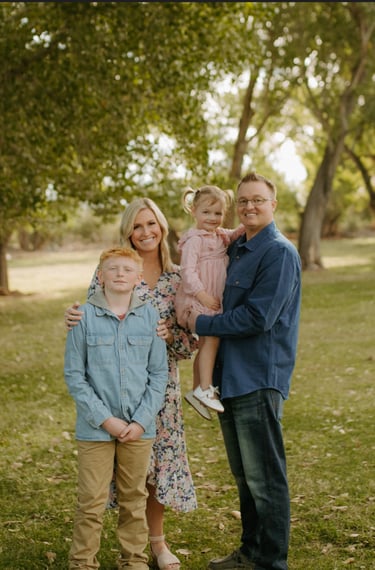 A happy family posing for an outdoor portrait in a grassy park surrounded by green trees.