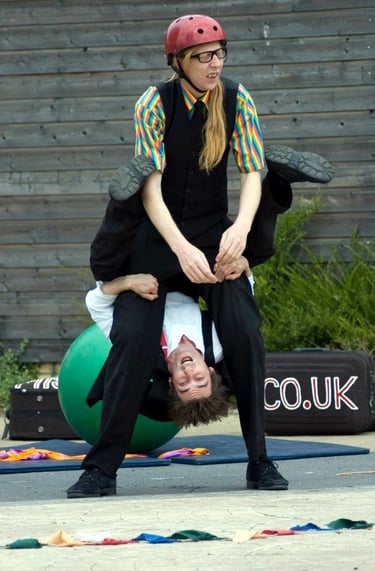 Two street performers in colorful costumes demonstrate a vertical acrobatic juggling balancing act. 