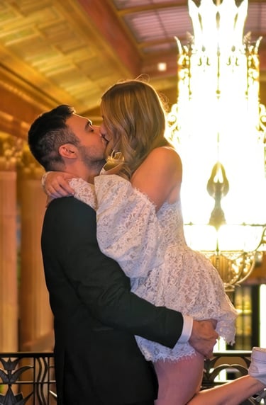 Couple embracing and kissing indoors with elegant chandelier lighting in the background, bride weari