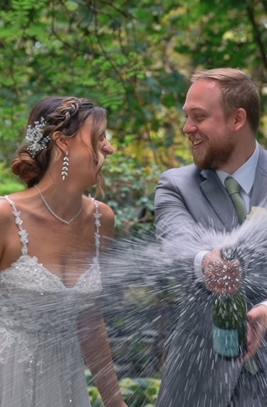 A bride and groom laugh together as the groom pops a champagne bottle, sending a spray of bubbles an