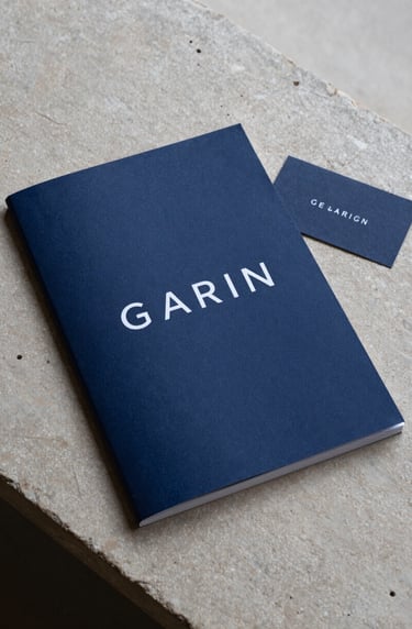 A top-down shot of an elegant exhibition catalog and a business card on a stone surface in a European / French gallery. The catalog features a dark navy blue cover with minimalist white typography. Clean, modern composition.
