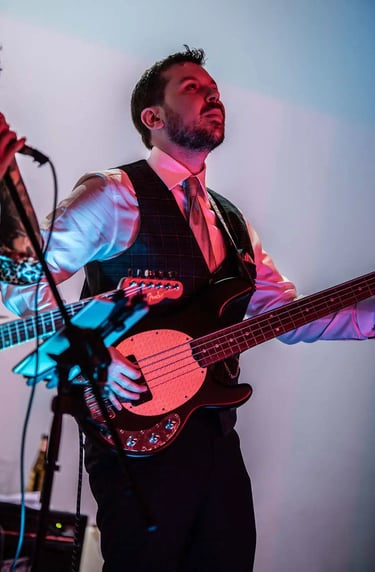 Groom plays a black electric bass guitar on stage with red and blue lighting.