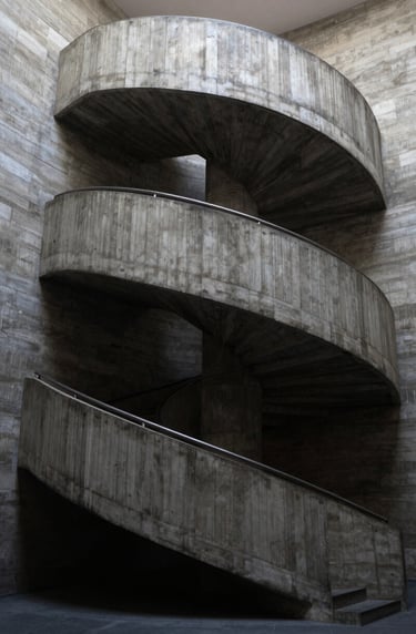 A vertical composition of a spiral concrete staircase in a South American / Brazilian art gallery. The lighting is soft but the shadows are deep black, emphasizing the sculptural quality of the architecture.