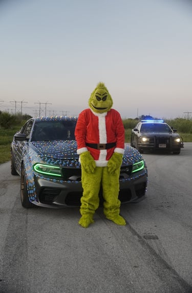 The Grinch in a Santa suit standing by a Dodge Charger with Christmas lights and a police car behind it.