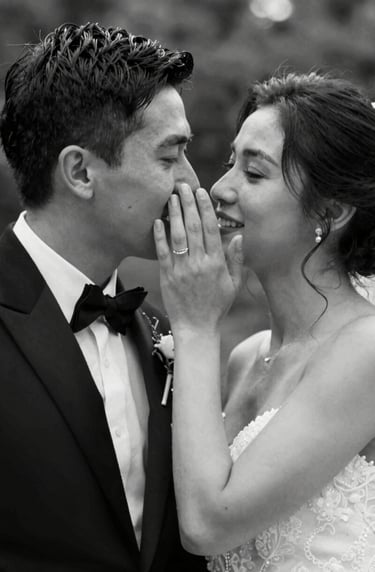 A black and white cinematic still (#20232B and #F7F5F0) showing a groom whispering to his bride. The composition is intimate and storytelling-focused, with elegant light grain texture.