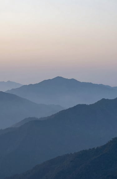A serene and clean landscape photograph of a misty mountain range at dawn. The colors transition from pale fog at the peaks to steel blue in the valleys. Minimalist composition focusing on natural scale and silence. International / Western landscape.