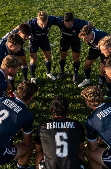 A cinematic overhead view of a team huddle on a green field. Sharp focus on the textures of jerseys in dark blue and black, dramatic low-angle evening sun, professional sports storytelling, Western / International.