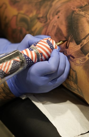 Professional tattoo artist in blue gloves using an American flag themed machine to ink a detailed design.