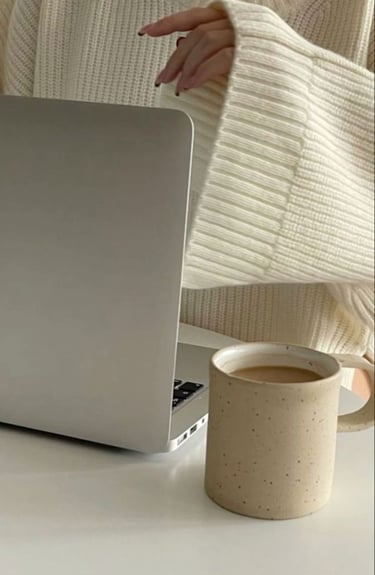 Woman in a cozy cream sweater working on a laptop with a coffee mug on the desk.