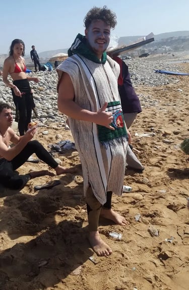Smiling man wearing a traditional striped djellaba and face paint on a sunny beach.