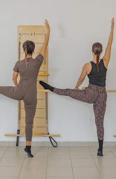 Two women performing a side leg raise stretch against the barre in a Pilates class.