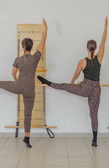 Two women performing a side leg raise stretch against the barre in a Pilates class.