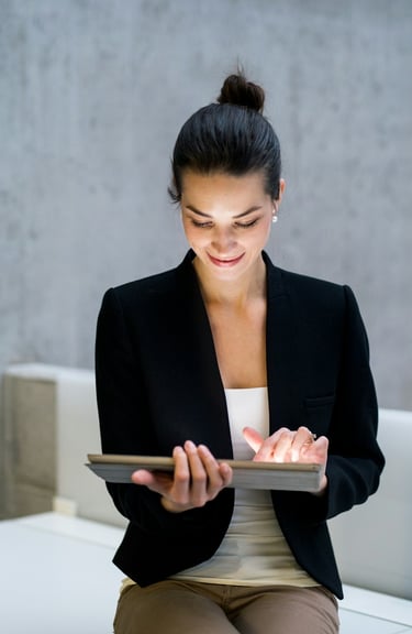A businesswoman in a black jacket, sitting on a desk, holding a tablet computer.