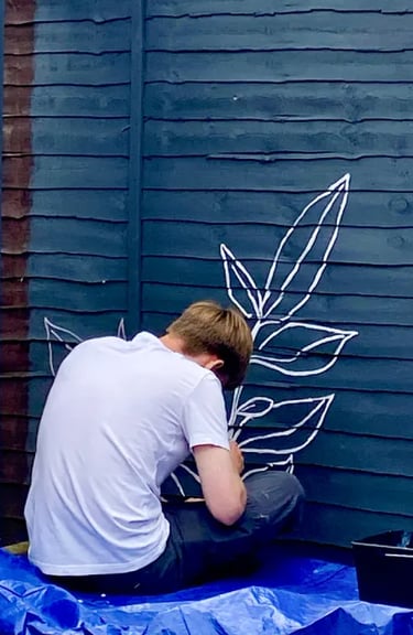 Muralist Byron sits on floor, painting graphic plant on grey wooden garden fence.