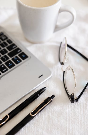 A laptop, pens, reading glasses, and coffee cup on a white table